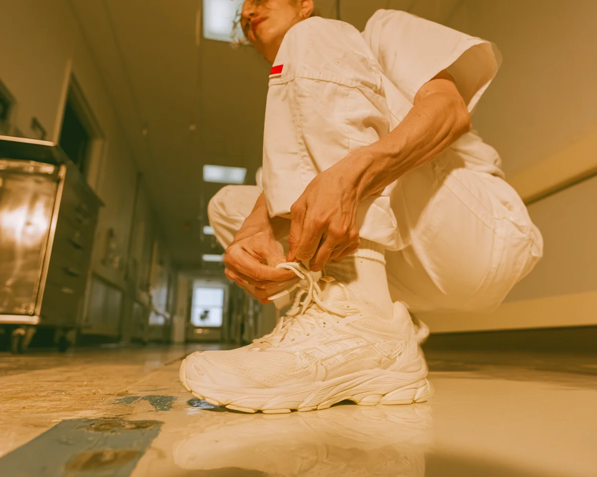 Someone in white work clothes lacing a cushioned shoe in a hospital corridor at the end of a shift
