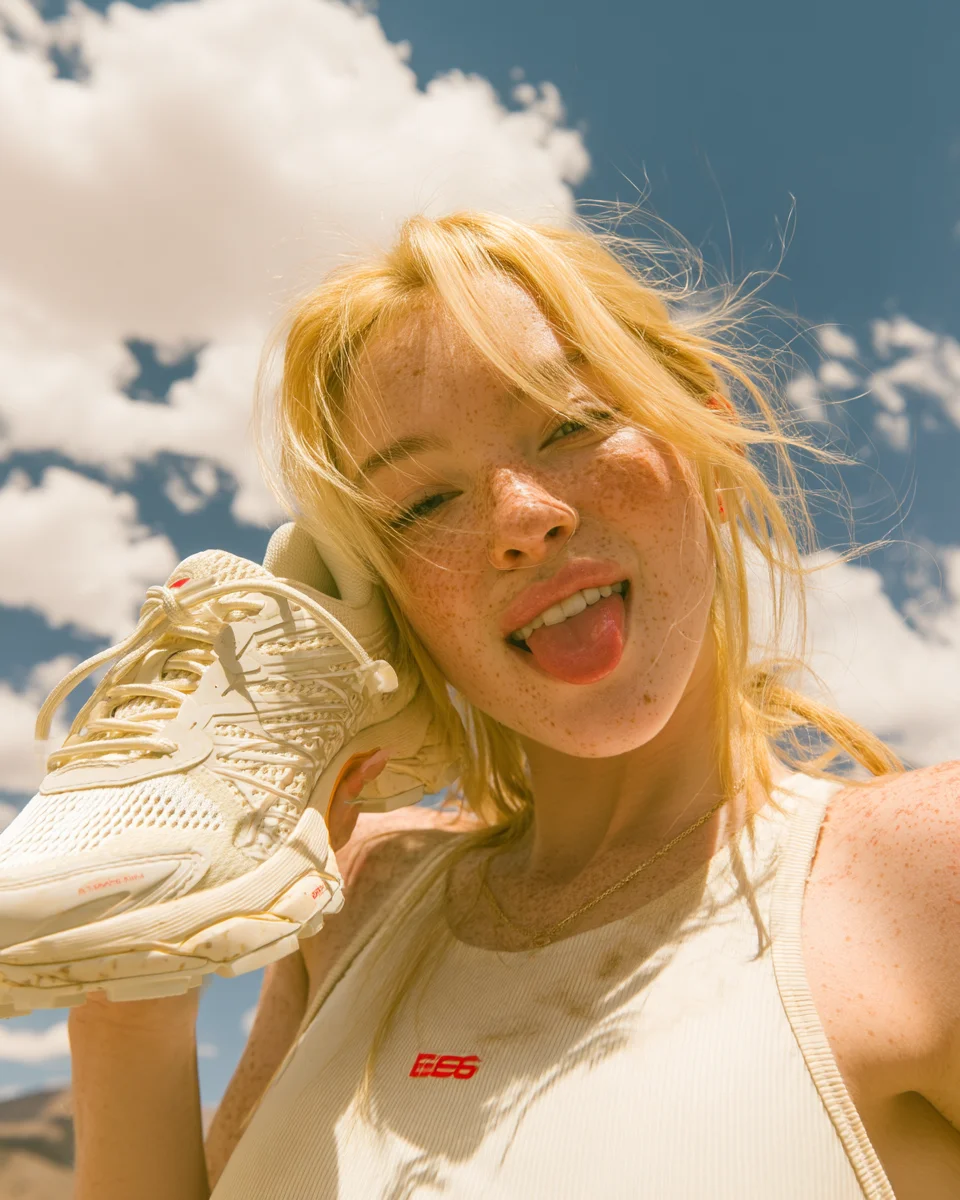 A runner laughing as she holds a cream trail shoe up beside her face against a bright Utah sky